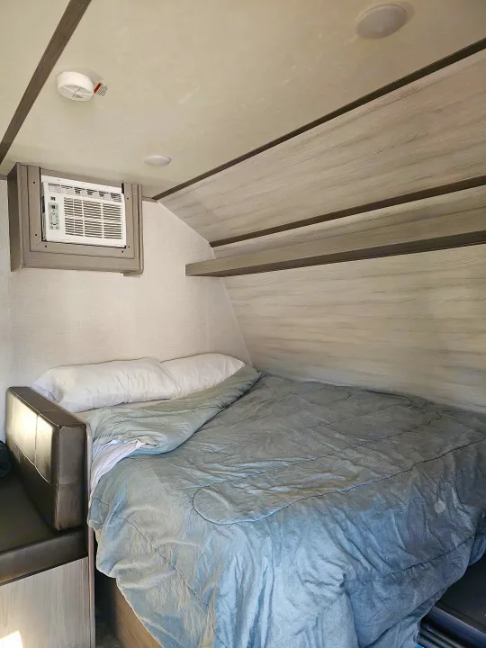 Compact RV rental bedroom showing a comfortable bed, window AC unit, and built-in shelving.. Dutchmen Coleman 17B 2023
