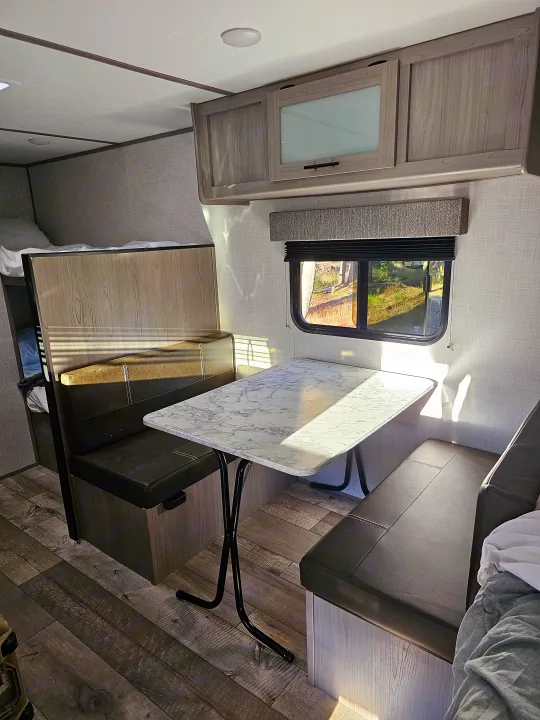 Modern RV rental interior: dinette with marble table and dark benches, upper bunk, bright window view.. Dutchmen Coleman 17B 2023