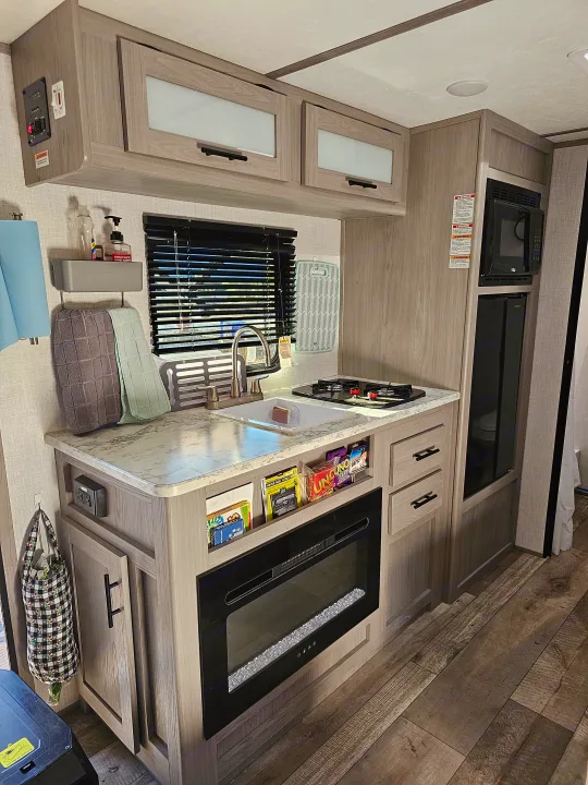 Inside this RV rental: a well-equipped kitchen with sink, stovetop, microwave, cabinets, and a cozy fireplace.. Dutchmen Coleman 17B 2023