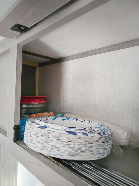 RV rental kitchen storage: open cabinet with stacks of paper plates, red bowls, and a Ziploc bag box.. Dutchmen Coleman 17B 2023