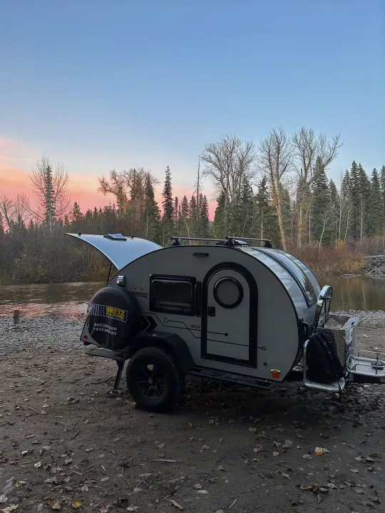 Light grey RV rental teardrop camper parked by a river at pink sunset, rear hatch open for adventure.. nuCamp T@G 2023
