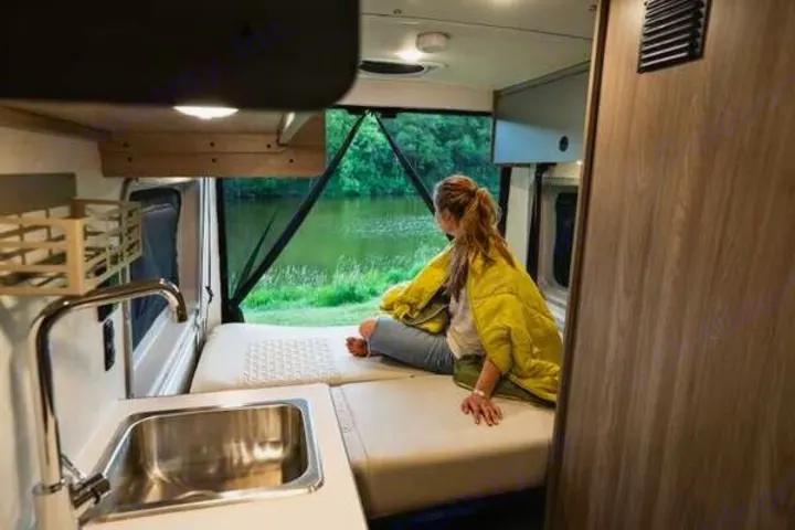 Relaxing RV rental moment: A woman on a bed looks out a large window at a serene lake, with a sink nearby.. Winnebago Solis 2023