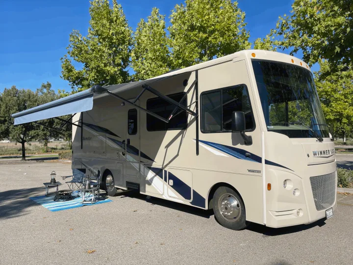 Spacious beige RV rental with extended awning, chairs, and mat on asphalt, perfect for outdoor leisure.. Winnebago Sunstar 2021