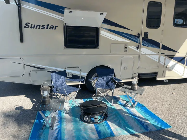 RV rental: Sunstar motorhome campsite with outdoor TV, two blue chairs, and a portable grill on a striped mat.. Winnebago Sunstar 2021