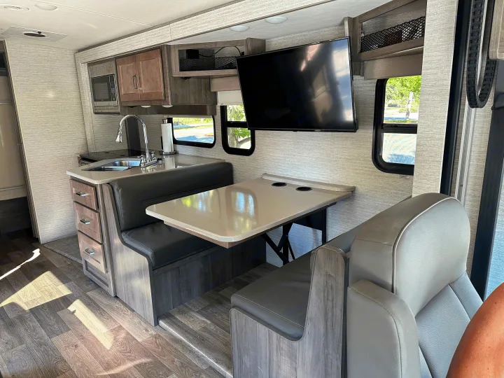 Inside a modern RV rental: compact kitchen with sink, built-in microwave, a dinette with TV, and wood-look flooring.. Winnebago Sunstar 2021