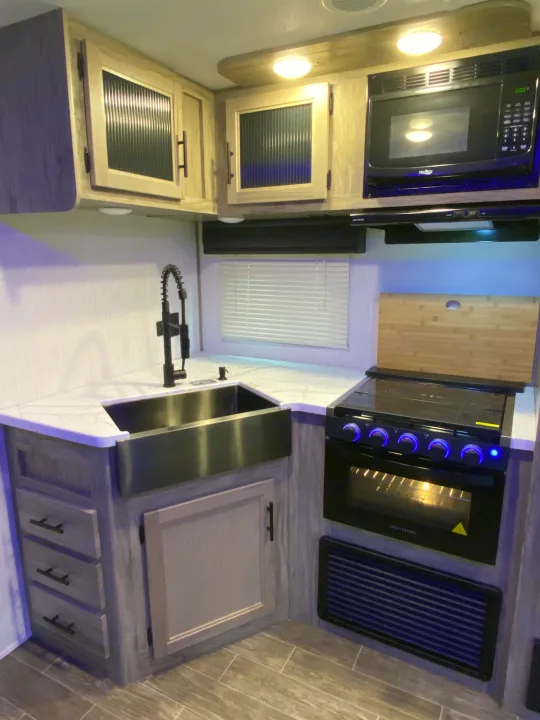 Modern RV rental kitchen featuring a stainless steel sink, black faucet, Greystone oven, microwave, and light wood cabinets.. Forest River Cherokee Alpha Wolf 2022