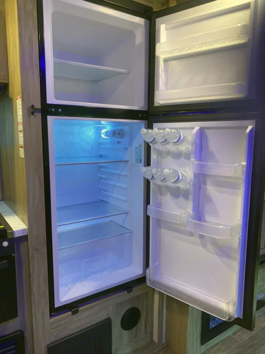 Empty and illuminated refrigerator inside an RV rental, showcasing clear shelves, a small freezer, and water bottles.. Forest River Cherokee Alpha Wolf 2022