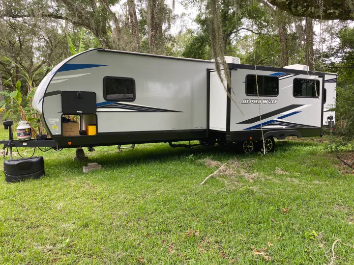 An "Alpha Wolf" travel trailer RV rental, grey with blue accents, parked on grass in a shaded, mossy campground setting.. Forest River Cherokee Alpha Wolf 2022