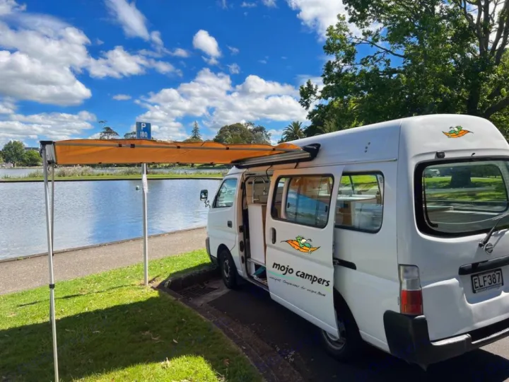 This white RV rental with an orange awning is parked lakeside, ready for outdoor adventure and camping.. Nissan Caravan 2005