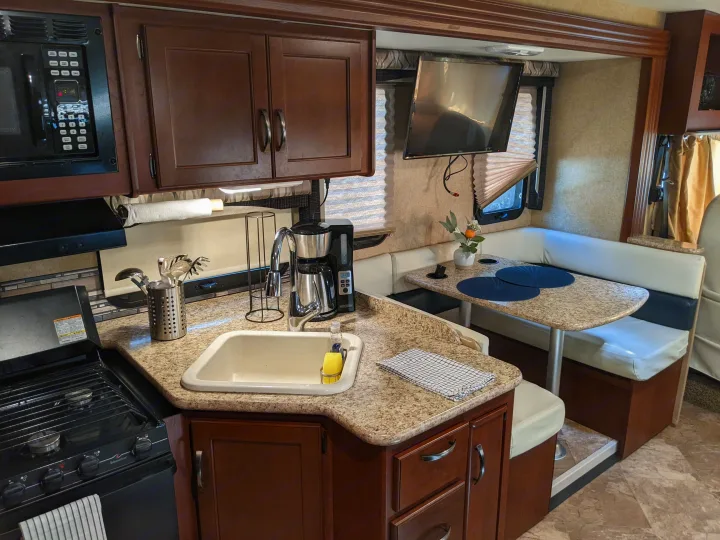 RV rental kitchen: microwave, stove, sink, coffee maker, with a dining booth and TV in the background.. Thor ACE 30.2 2015