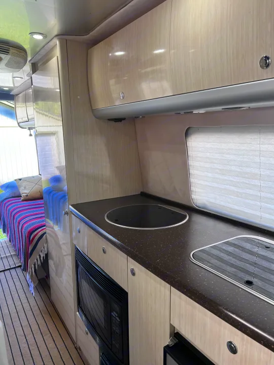 Step inside this RV rental! Modern interior with light wood cabinets, dark countertops, a built-in microwave, and a cozy bed.. Airstream Interstate 2012