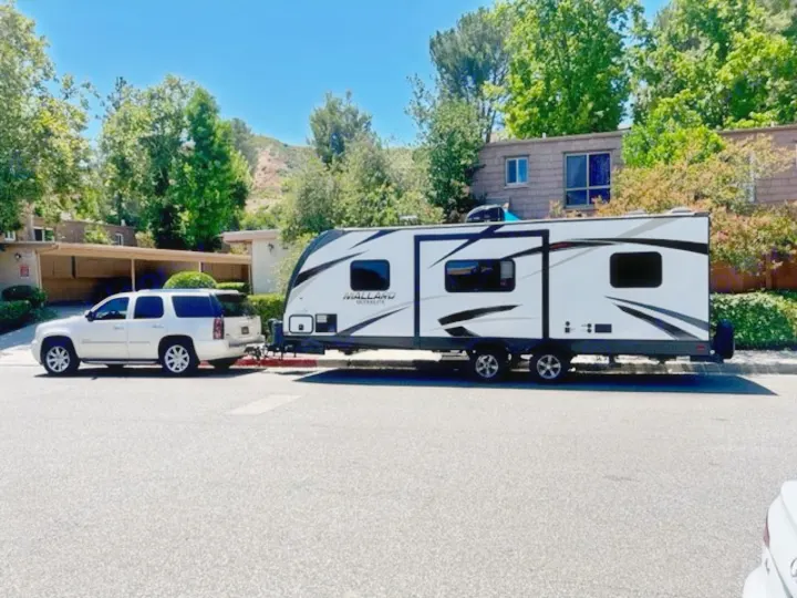 White SUV towing a Mallard Ultralite RV rental, parked on a sunny street.. Heartland RVs Mallard 2018