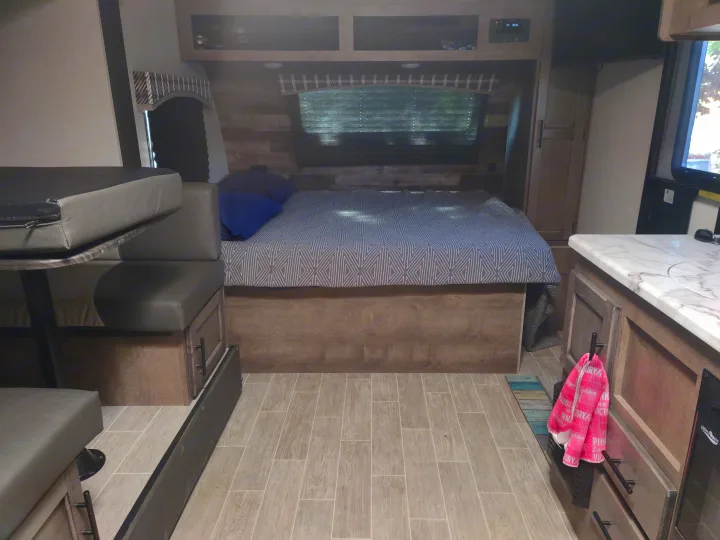 Interior view of an RV rental, featuring a comfortable bed, dinette, and kitchen space with light wood tones.. Sonic Lite 2021
