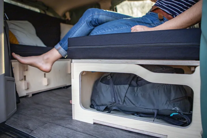 RV rental: Camper bed with integrated plywood storage holding a backpack, and a person relaxing.. Chrysler Grand Caravan 2025
