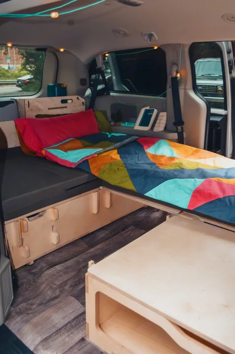 Bright & functional RV rental: comfy bed with a colorful quilt, smart wooden storage, and string lights in a van conversion.. Chrysler Grand Caravan 2022