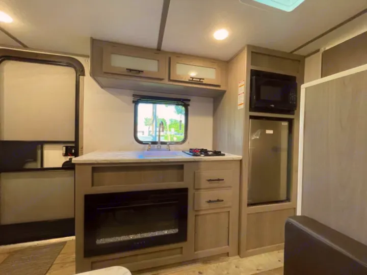 RV rental kitchen: Features a sink, 2-burner stovetop, microwave, compact fridge, and electric fireplace in modern cabinetry.. Coleman Lantern 17B 2024