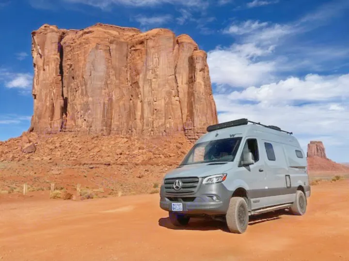 Grey RV rental van on red dirt road. Towering desert buttes under blue sky. Your next adventure awaits!. Winnebago Revel 2022