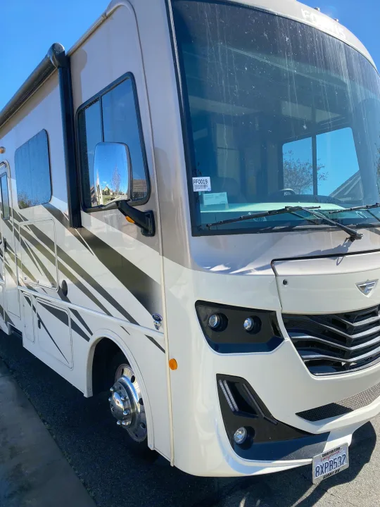 Front-side view of a white and tan RV rental featuring black striped graphics, chrome wheels, and a visible California license plate.. Fleetwood Fortis 2021
