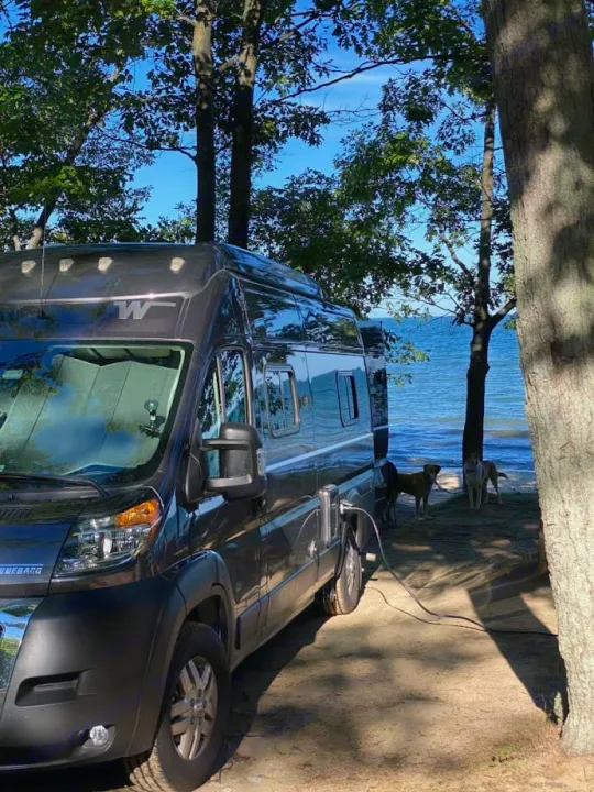 An RV rental, a dark grey camper van, parked lakeside with two dogs by calm blue water & trees. Perfect Winnebago camping trip.. Winnebago Solis 2023