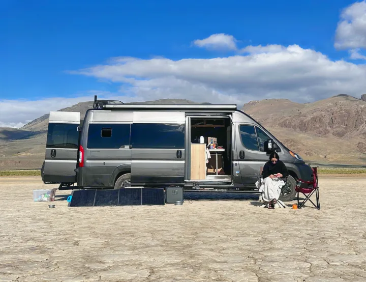 A grey RV rental van, doors open, sits on a cracked dry lake bed. A person relaxes outside, using solar panels for off-grid power.. Airstream Airstream Class B 2023