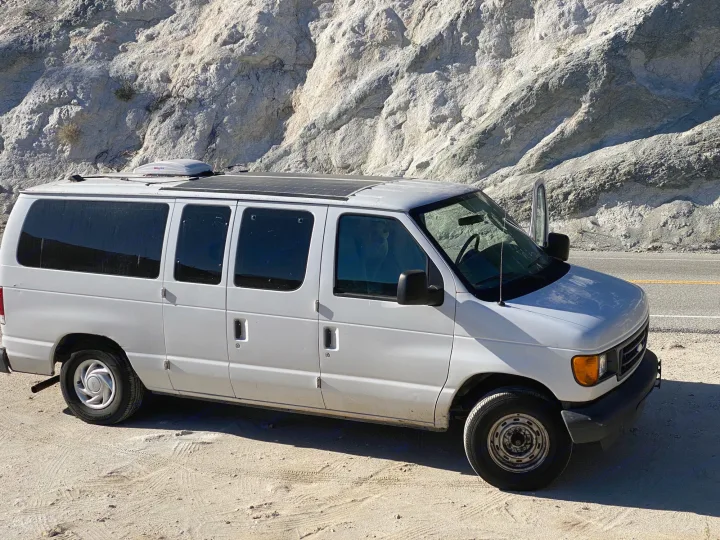 Converted white Ford van with solar panels, parked by mountains. Great RV rental for off-grid travel.. Ford Econoline 150 2003