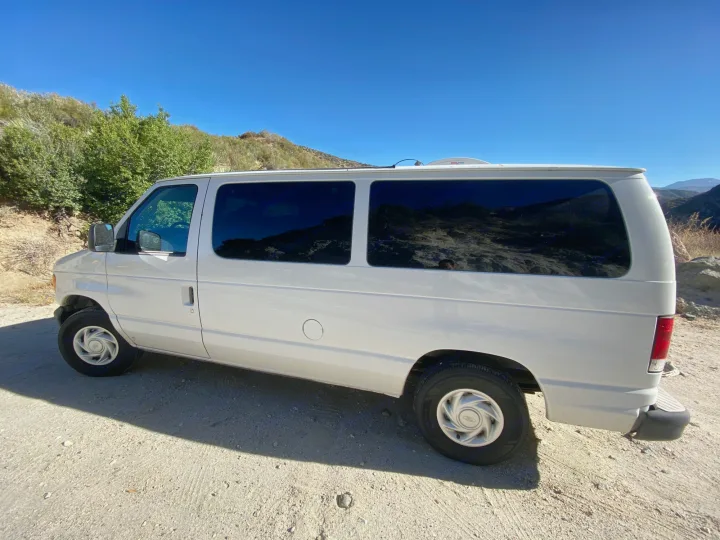 White van, an RV rental, parked on a sunny dirt road amidst green mountains under a clear blue sky.. Ford Econoline 150 2003