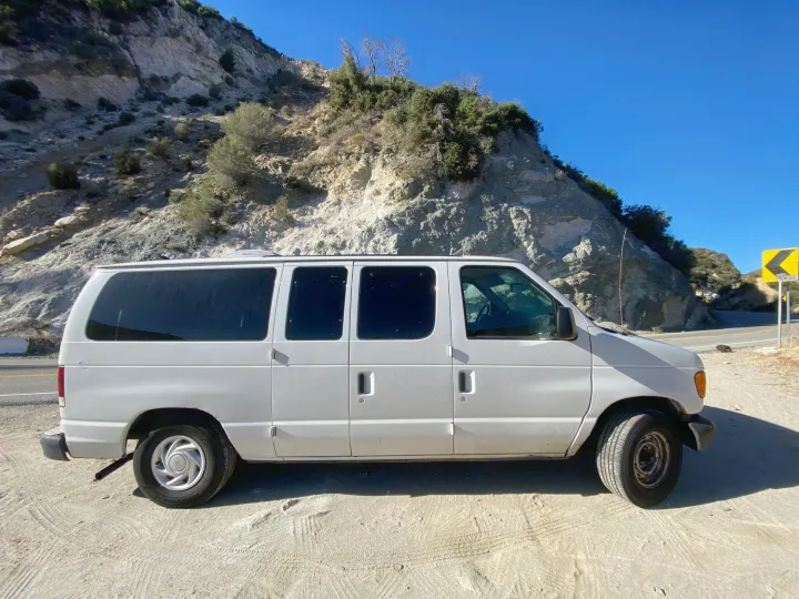 White passenger van, an ideal RV rental, parked on a dirt shoulder against a rugged mountain landscape under a clear sky.. Ford Econoline 150 2003