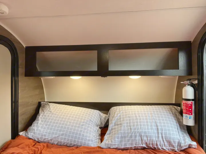 RV rental interior showing a bed with pillows, overhead cabinets, reading lights, and a visible fire extinguisher for safety.. Braxton Creek Free Solo 2022