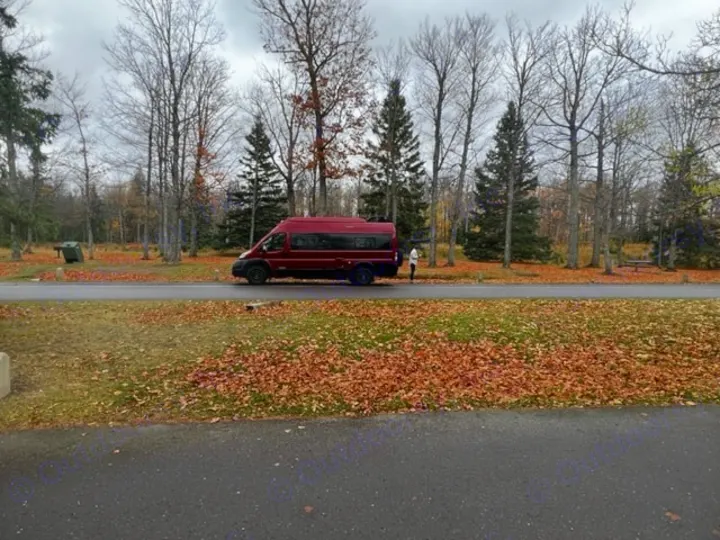 Autumn RV rental: A red camper van is parked on a path in a park, with a person nearby and fall leaves covering the ground under a cloudy sky.. Winnebago Solis 2021
