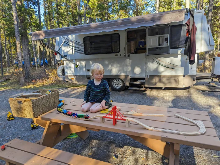 Young child plays with trains on a picnic table beside an RV rental in a wooded campsite, enjoying outdoor family fun.. Hi Lo 2307c 2007