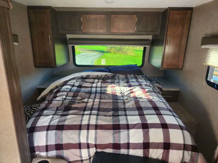 Cozy RV rental bedroom featuring plaid bedding, overhead cabinets, and a scenic window view. Ideal for travel trailer getaways.. Forest River Rockwood Mini Lite 2019