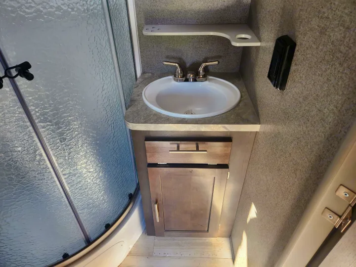 This compact RV rental bathroom features a white sink, modern wood-grain vanity, and a textured glass shower.. Forest River Rockwood Mini Lite 2019