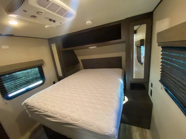 Inside an RV rental: a compact bedroom with a white bed, overhead AC, built-in storage, and window blinds.. Grand Design Transcend 2021