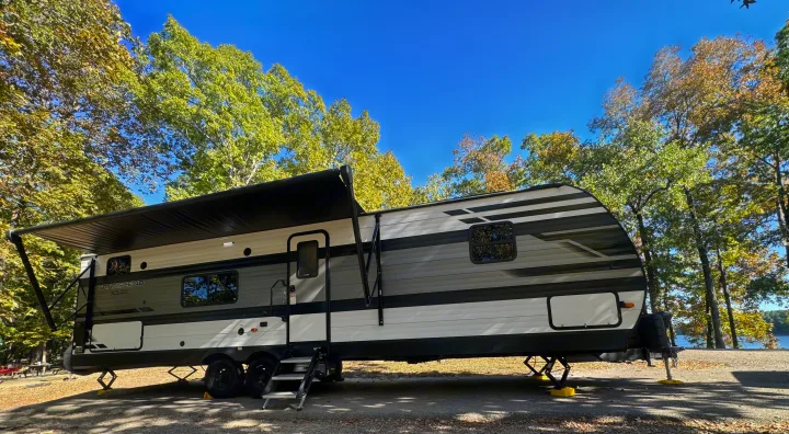 White & gray RV rental with awning extended at a sunny lakeside campsite among trees.. Grand Design Transcend 2021