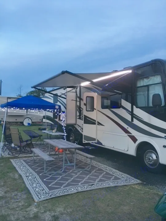 Cozy RV rental campsite at dusk, featuring a motorhome with illuminated awning, outdoor rug, picnic area, and blue canopy.. Forest River Coachmen Pursuit 2017