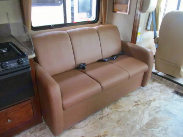 RV rental: Light brown sofa with seatbelts, adjacent to a compact kitchen featuring a stovetop and counter.. Forest River Coachmen Pursuit 2017