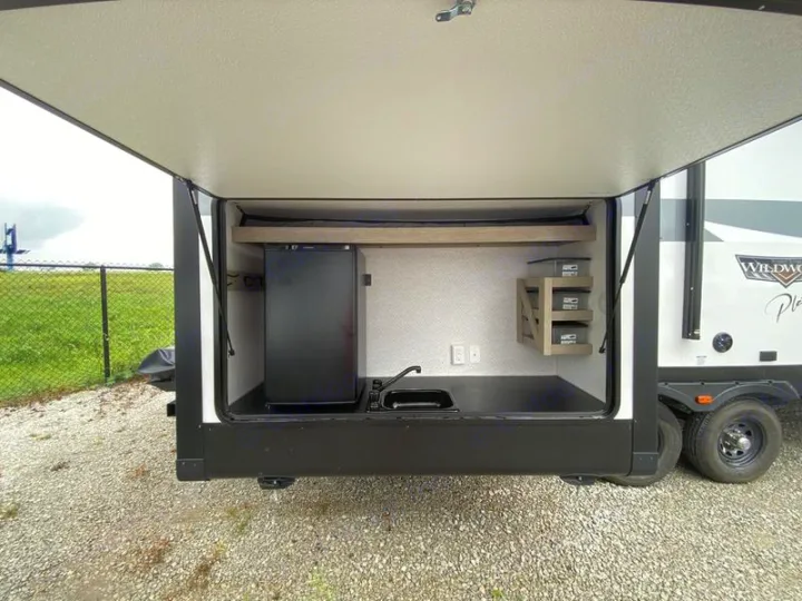 An open RV side compartment showcases an outdoor kitchen with a mini-fridge, sink, and shelving, perfect for rv rental.. Forest River 31KQBTS 2024