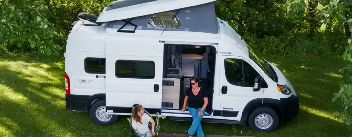 Two women relax by a white camper van RV rental with an extended pop-top roof, enjoying the outdoors.. Winnebago Solis 2022
