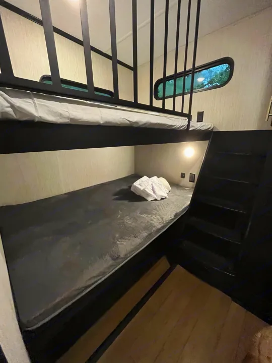Cozy RV rental bunk beds feature dark grey mattresses, a window, and a ladder for comfortable adventures.. Forest River Wildcat 2022