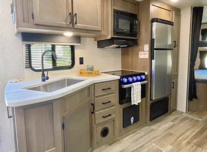 The well-equipped kitchen of an RV rental, showing a sink, microwave, stove, and refrigerator, with wood-look cabinets and flooring.. Starcraft Autumn Ridge 2023