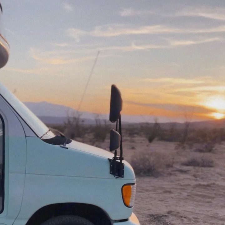 RV rental van in a desert sunset. Light blue camper van with prominent mirrors faces arid landscape and distant mountains under a warm sky.. Ford E450 1999