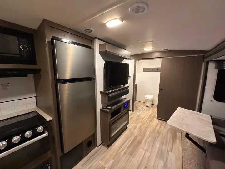 Inside a modern RV rental: full kitchen with stainless fridge, TV entertainment center, and bathroom entrance. Perfect compact living.. Cruiser RV MPG Ultra Lite 2022