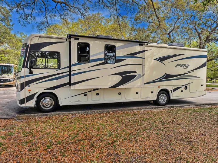 Cream-colored RV with black/gray decals, extended slide-out, parked on asphalt with autumn leaves. Perfect for an RV rental trip.. FR3 FR3 Motorhome 2020