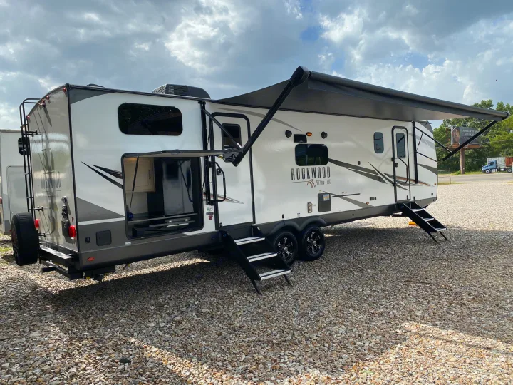 Bright white and gray Rockwood RV rental travel trailer on gravel, featuring an extended awning and outdoor kitchen.. Forest River Rockwood 2023
