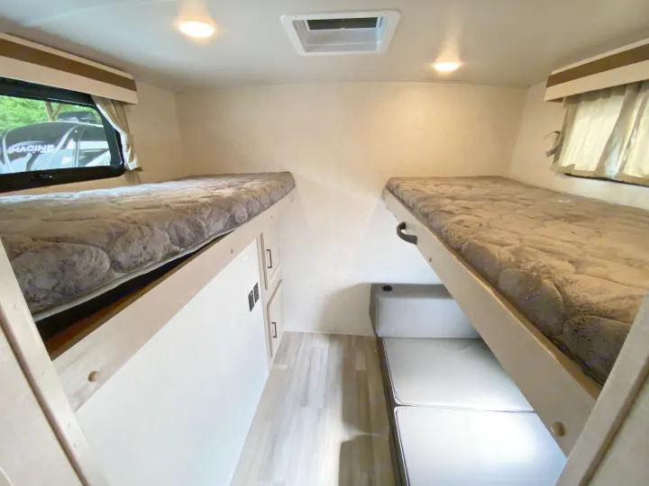 Inside an RV rental, showing two bunk beds with soft gray bedding. One bunk folds up above a seating area, ideal for compact living.. Forest River Rockwood 2023