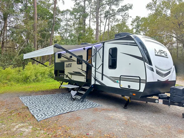 A white RV rental parked at a campsite, with its awning extended and a patterned mat laid out.. Cruiser RV MPG Ultra Lite 2022