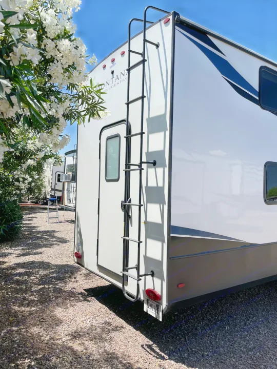 Explore a Montana High Country RV rental: a white fifth wheel with a rear ladder, nestled among white flowers on gravel.. Keystone RV Montana High Country 2023