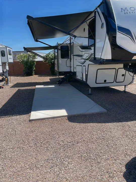 This white & black Montana fifth-wheel RV rental, with extended awnings, rests on a gravel lot next to a concrete patio.. Keystone RV Montana High Country 2023