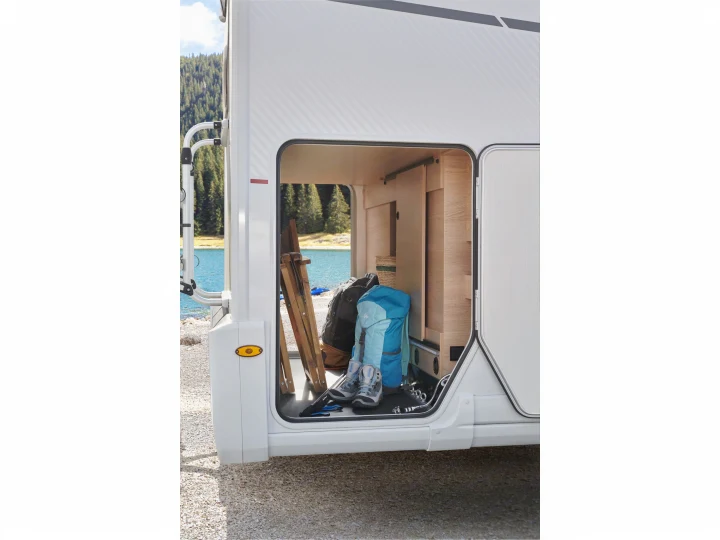 RV rental's side storage is open, revealing hiking gear: boots, backpacks, and a folding chair, with a lake in the background.. PILOTE A696G Atlas 2025