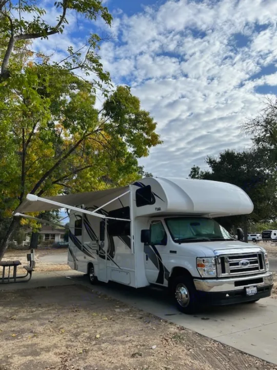A white RV rental, a Class C motorhome, with its awning extended, parked at a treed campsite under a cloudy sky.. Thor Four Winds 2022